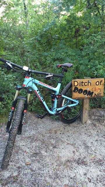 Trek Fuel EX 8 29: A mountain bike parked next to a wooden sign that reads "Ditch of Doom." The bike features a light blue frame and is positioned on a dirt path surrounded by dense greenery and trees. The sign is hand-painted and adds an adventurous feel to the scene.