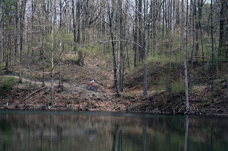 A scenic view of a forested area with a cyclist biking along a dirt path beside a calm lake, surrounded by trees in early spring. The landscape is characterized by bare branches and emerging greenery, reflecting off the water's surface. Griffin Bike Park mountain bike trail.