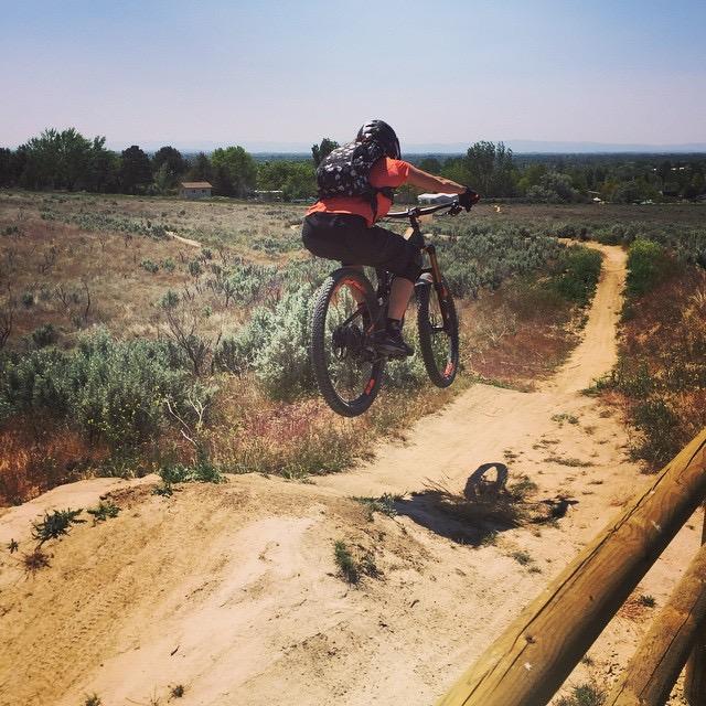 A mountain biker in a bright orange shirt jumps off a dirt ramp, catching mid-air above a sandy bike trail surrounded by shrubs and trees. The sky is clear and blue, highlighting the outdoor setting. Eagle Bike Park mountain bike trail.