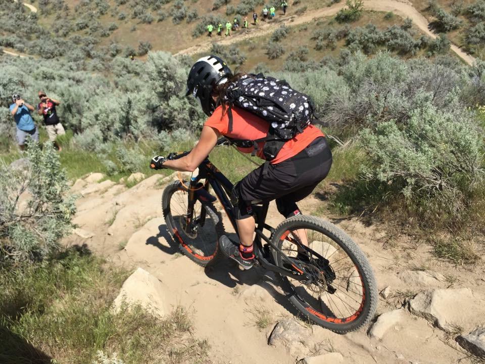 A mountain biker in an orange shirt and black shorts descends a rocky trail surrounded by greenery. The biker is wearing a helmet and a backpack, focused on navigating the uneven terrain. In the background, a group of people in bright green shirts can be seen hiking up the trail. Eagle Bike Park mountain bike trail.