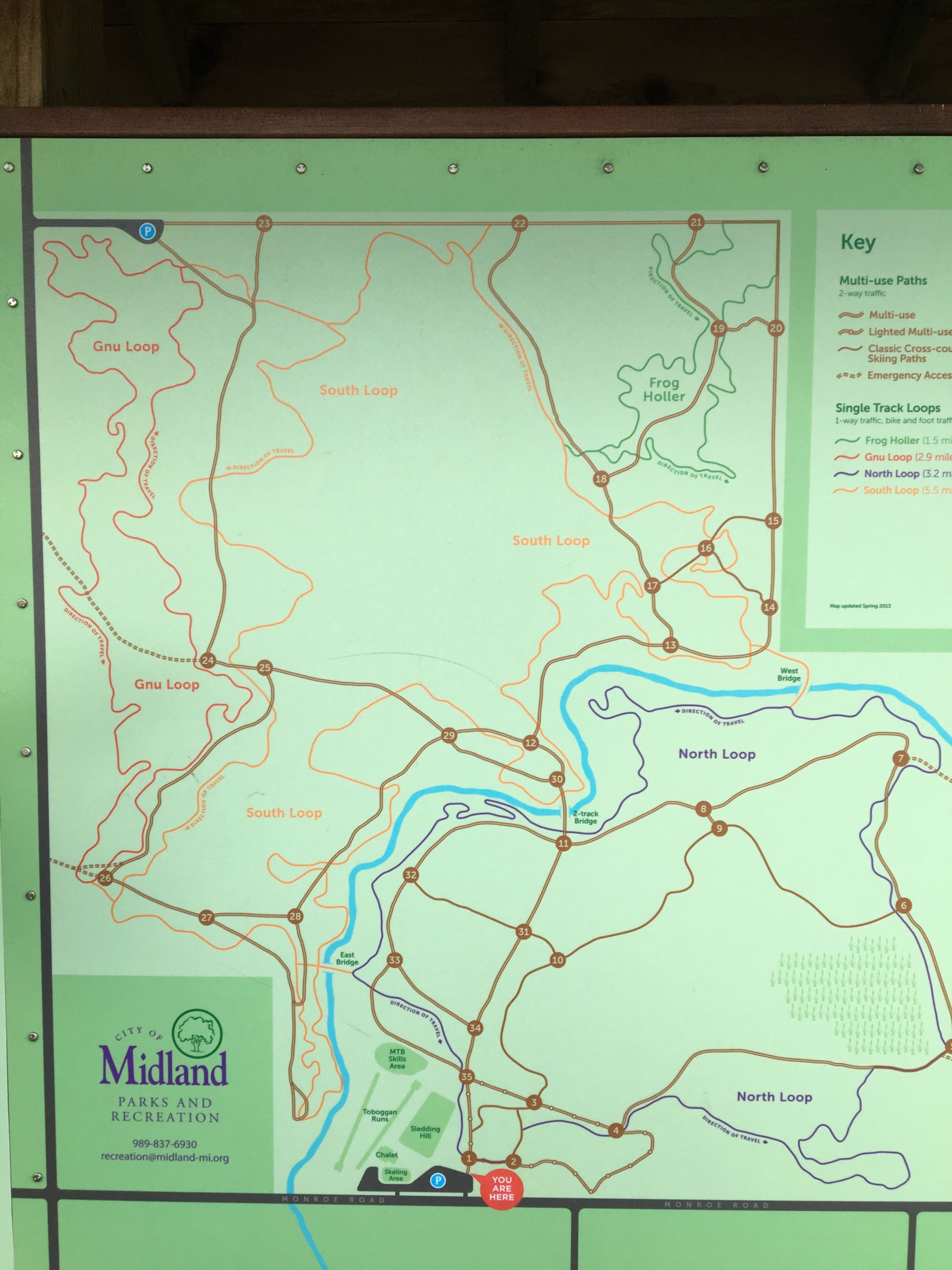 A colorful trail map showing various loops and paths in a park, including the Gnu Loop, South Loop, and North Loop. The map features labeled trails, key for multi-use and single track paths, and location indicators. A "You Are Here" marker is present, along with contact information for Midland Parks and Recreation. Midland City Forest mountain bike trail.