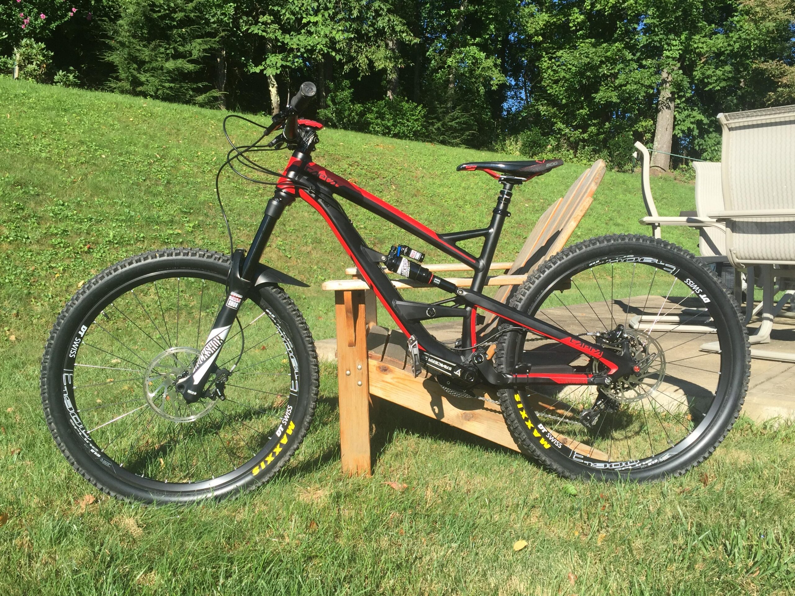 YT Capra: A black and red mountain bike is leaning against a wooden chair in a grassy outdoor setting. The bike features large, knobby tires and a sturdy frame, suggesting it's designed for off-road use. In the background, a green lawn and patio furniture are visible under bright sunlight.