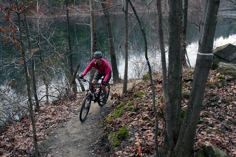 A mountain biker wearing a pink long-sleeve shirt and a helmet rides along a narrow trail by a lake, surrounded by trees that are bare of leaves, with patches of fallen leaves on the ground. The water reflects the landscape, creating a serene outdoor scene. Griffin Bike Park mountain bike trail.