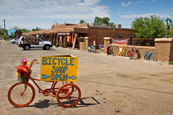 A vibrant bicycle shop with a colorful sign that reads "Bicycle Shop Open" displayed on an orange bike adorned with flowers. The shop's adobe-style building is in the background, along with various bicycles in cheerful colors lined up outside. The scene is set on a sunny day with a clear blue sky.