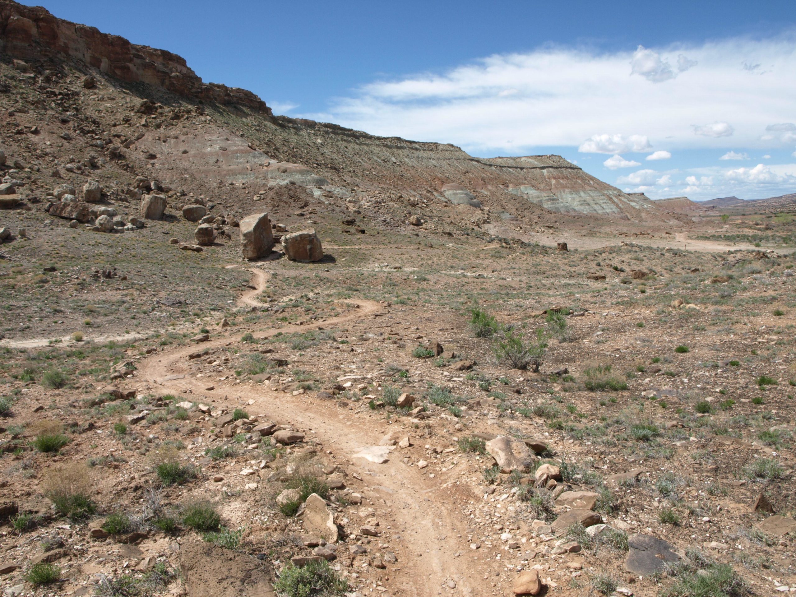 A winding dirt path leads through a dry, rocky landscape with sparse vegetation and large boulders, set against a backdrop of a blue sky with scattered clouds. The terrain features sloping hills and layered rock formations in the distance. Chilkoot Pass mountain bike trail.