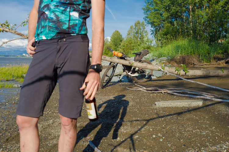 A person wearing a colorful, palm-patterned shirt and shorts stands by a beach, holding a bottle in one hand. In the background, a mountain bike leans against a fallen tree, with a scenic view of greenery and calm water. The sunlight casts shadows on the sandy ground.