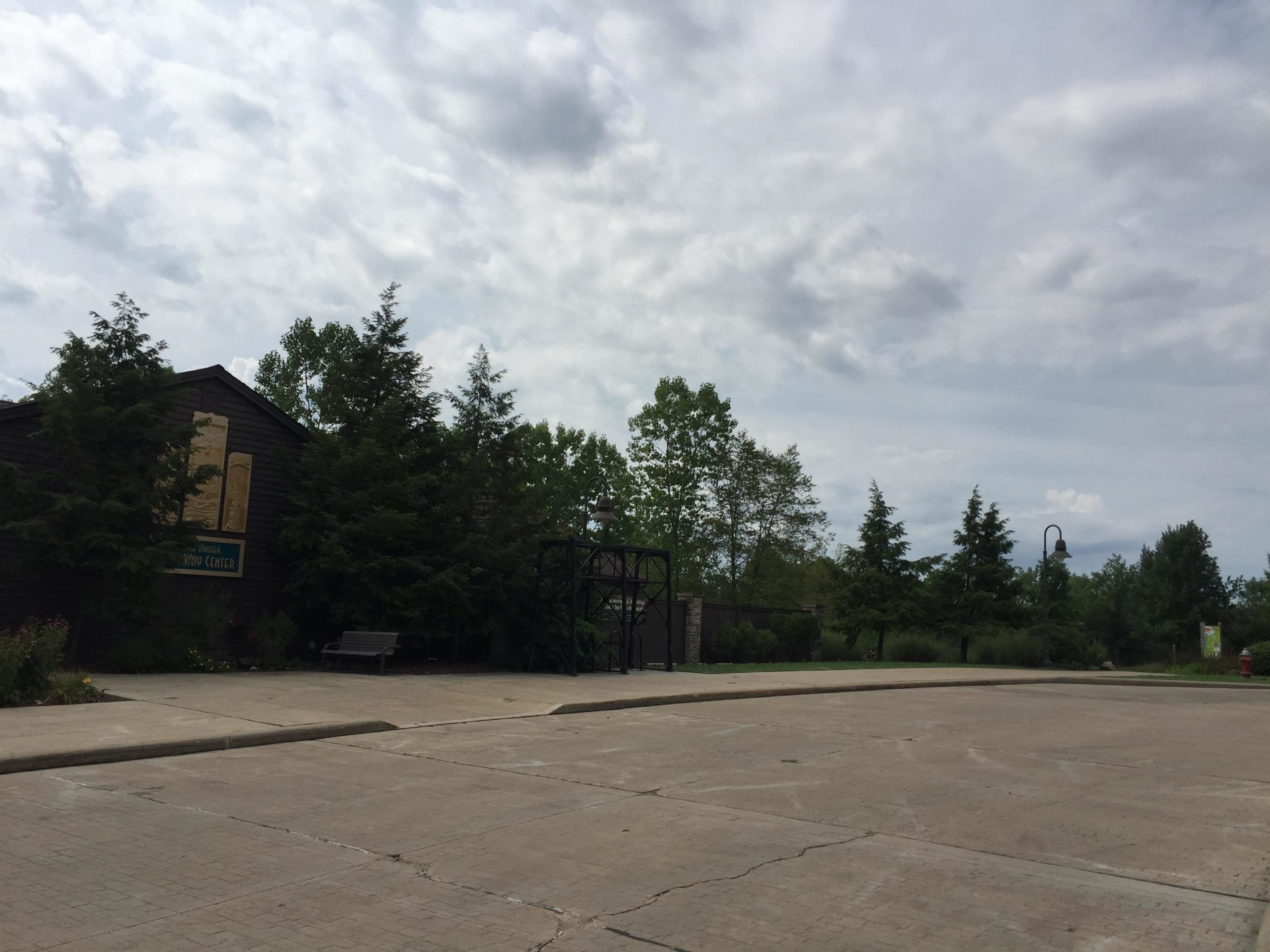 A view of a paved area in front of a nature center building surrounded by trees under a cloudy sky. There is a bench nearby and a light post in the background. The scene conveys a peaceful outdoor setting. Ohio And Erie Canal mountain bike trail.