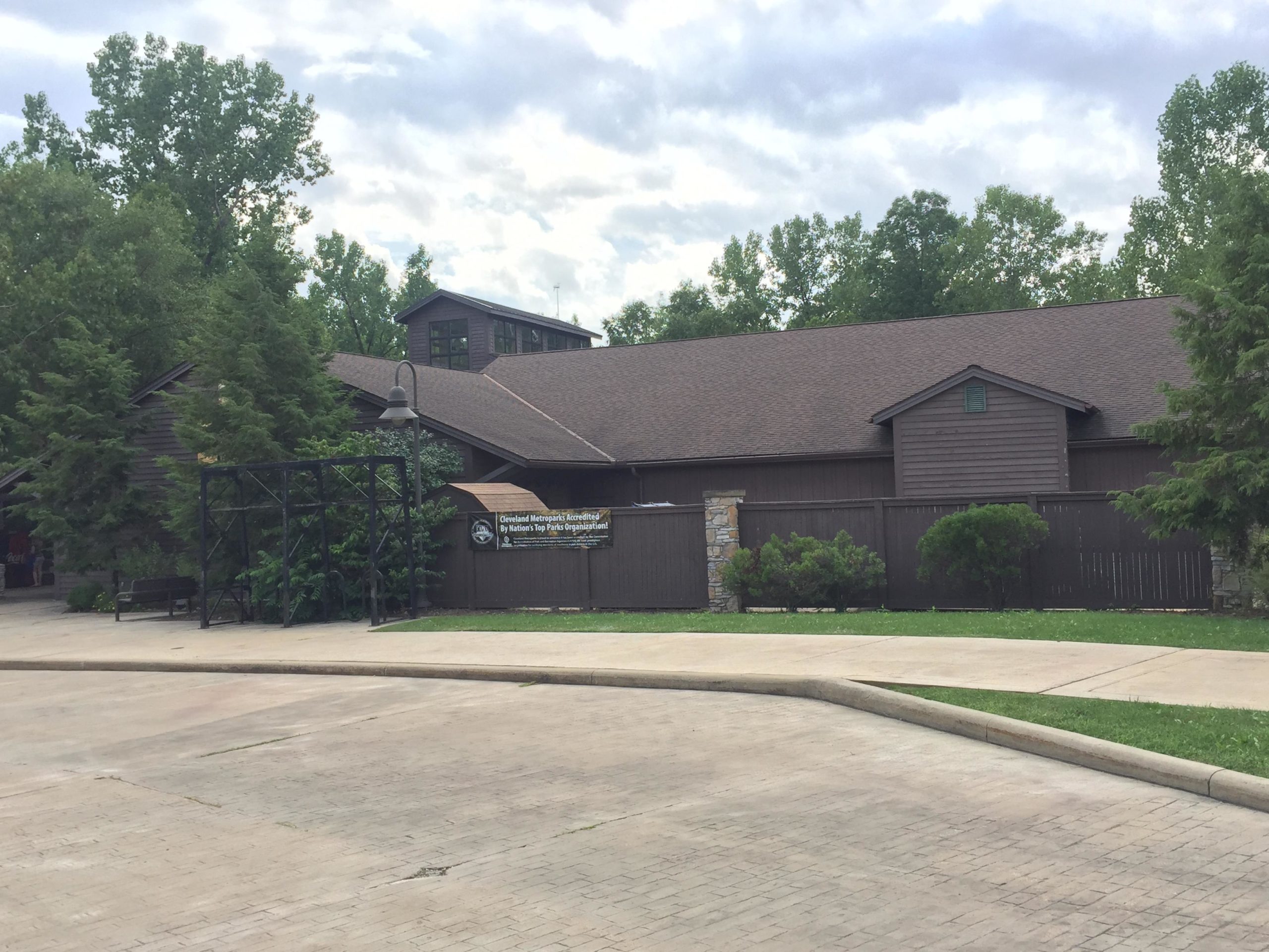 Exterior view of a brown wooden building surrounded by trees, with a sign reading "Cleveland Metroparks Accredited by Nation's Top Parks Organization." The pathway in front is paved, and there are shrubs and benches visible. The sky is partly cloudy. Ohio And Erie Canal mountain bike trail.