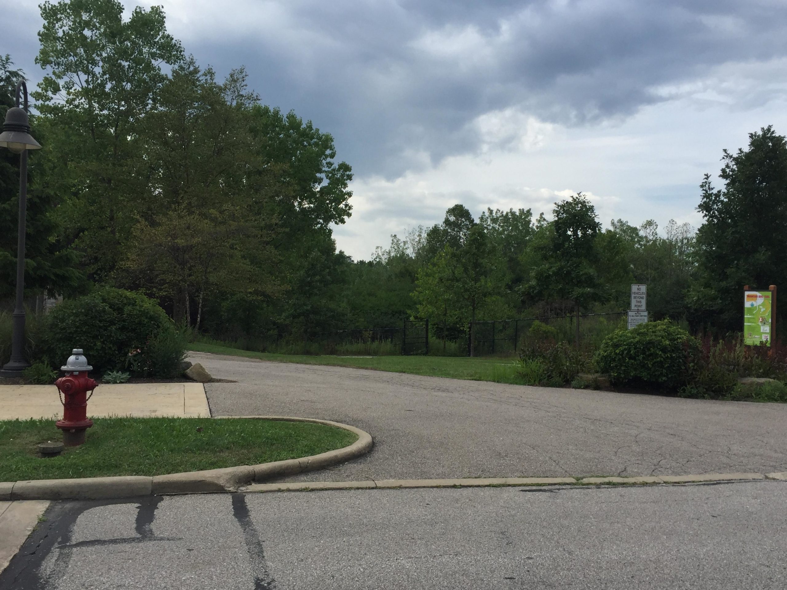 A quiet park entrance featuring a paved path leading into a wooded area, with trees on either side and a cloudy sky above. A red fire hydrant is positioned on the left, next to a grassy area, while a sign indicating park regulations is visible on the right. Ohio And Erie Canal mountain bike trail.