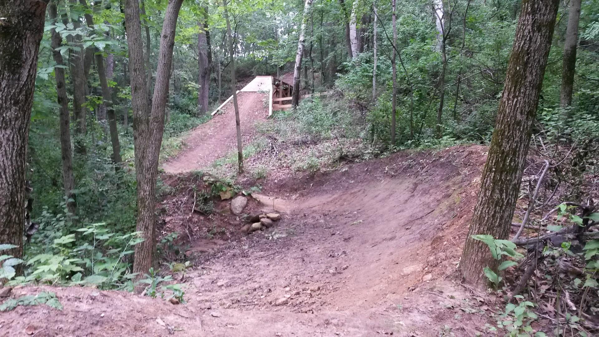A dirt bike jump ramp situated in a wooded area, surrounded by trees and greenery. The ramp leads up to a dirt trail that winds through the forest, with rocks and soil visible along the path. The scene captures the natural setting ideal for outdoor biking activities. Quarry Ridge Park mountain bike trail.