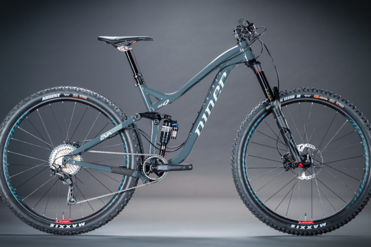A modern mountain bike in a sleek gray color with black and turquoise accents, featuring front and rear suspension, wide tires, and a compact frame design. The bike displays detailed components including the gearing system and disc brakes, set against a gradient background.