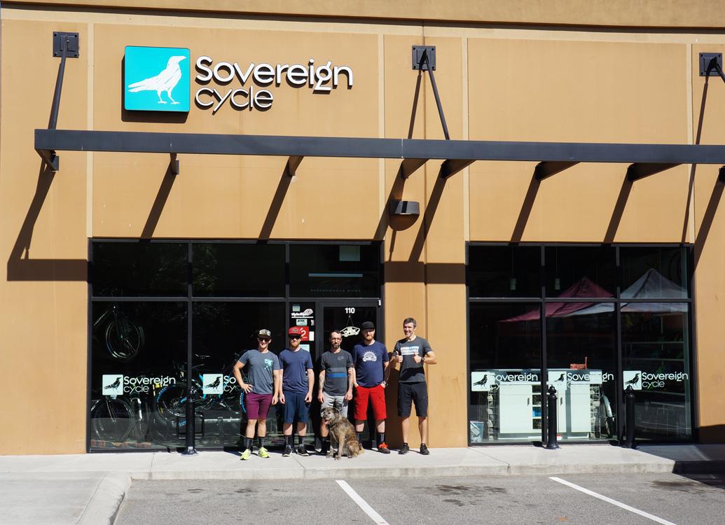 Group of five individuals standing outside the Sovereign Cycle store, with a dog present. The store features a prominent sign and large windows displaying bicycles inside. The people are casually dressed, and it appears to be a sunny day.