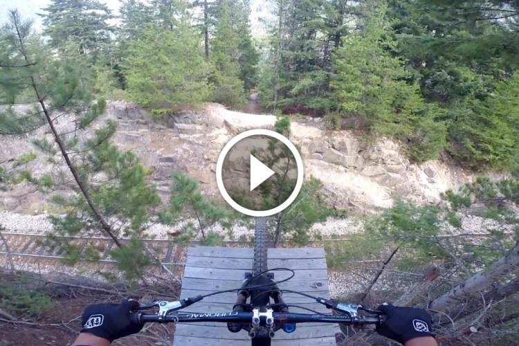 A mountain biker's view from a wooden platform, overlooking a rocky terrain and trees, with a play button overlay indicating a video.
