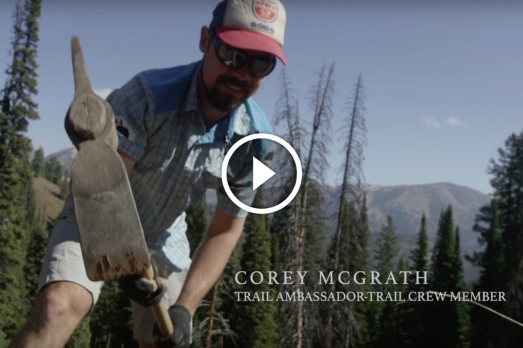 A person wearing sunglasses and a cap is working outdoors using a shovel in a forested area. In the background, there are trees and mountains, indicating a natural setting. Text at the bottom of the image identifies the person as a trail ambassador and crew member.