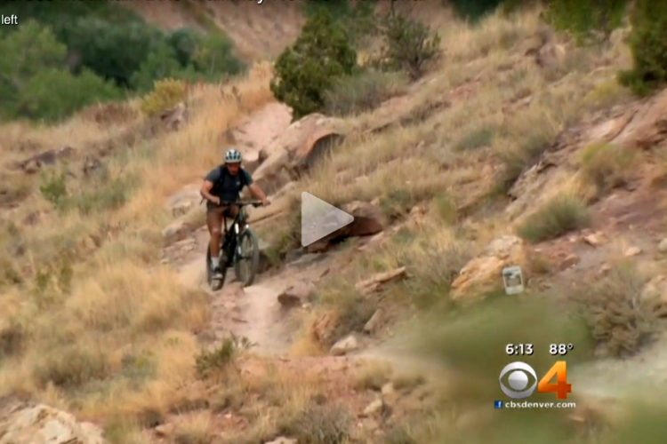 A person riding a mountain bike along a rocky trail surrounded by dry grass and shrubs in a hilly landscape.