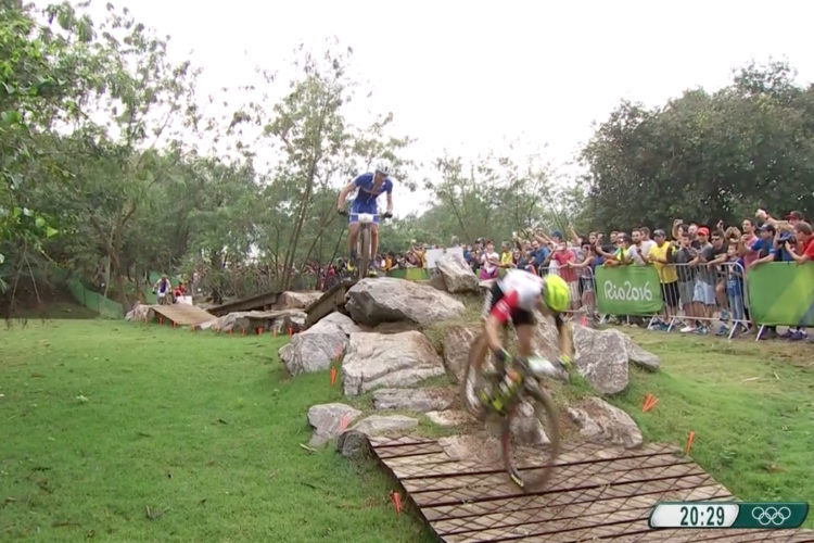 A mountain biker navigates a rocky terrain during a race, while a competitor jumps off a wooden ramp in the background. A crowd of spectators cheers on the racers, with trees providing a natural backdrop. The image captures the excitement and intensity of competitive mountain biking.
