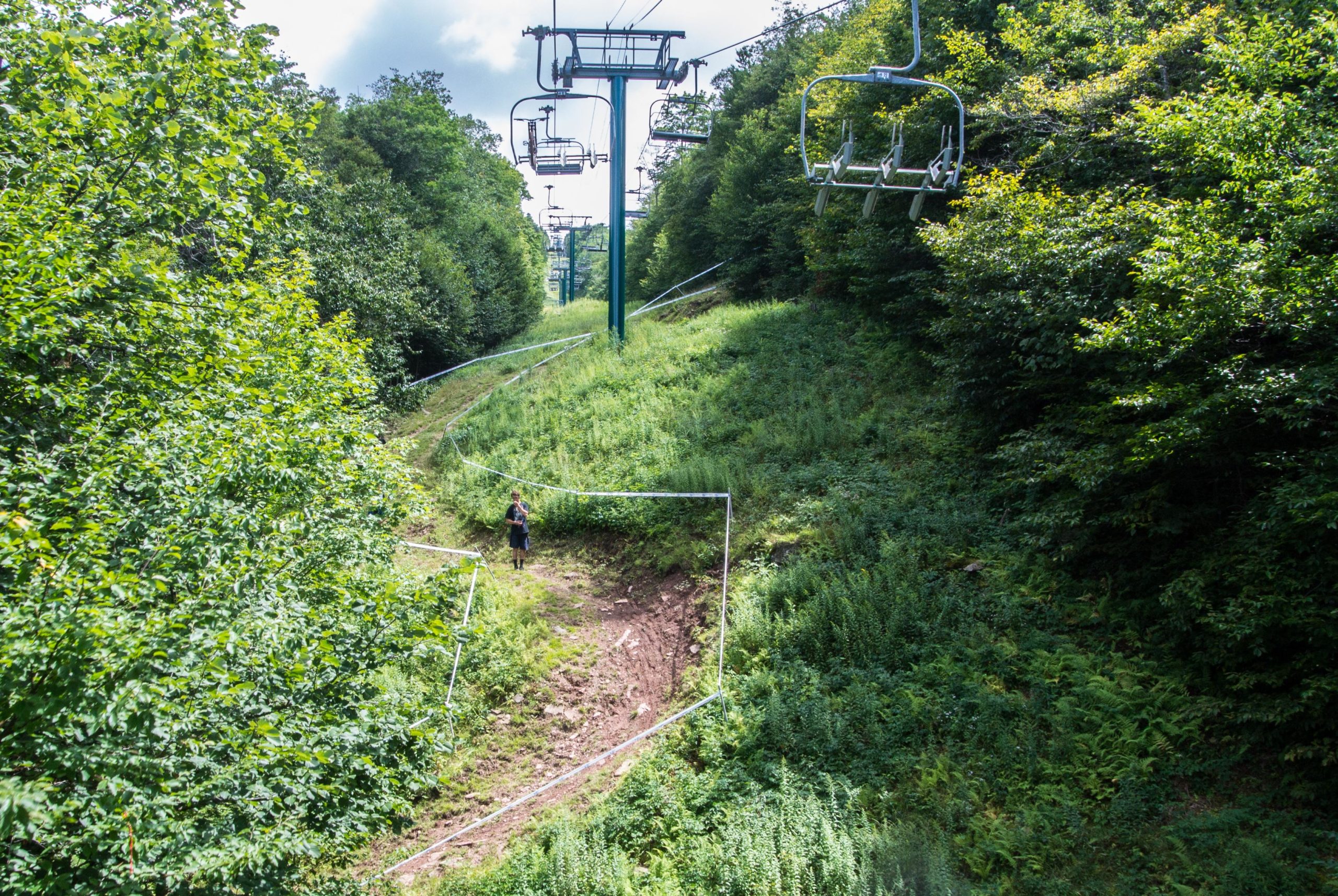 A scenic view of a ski slope surrounded by lush greenery, with a single person standing on a dirt path. The background features ski lift chairs and towers ascending the hillside, while the terrain is marked by rolled plastic fencing along the path. The sky is partly cloudy, creating a bright, natural atmosphere. Snowshoe Bike Park mountain bike trail.