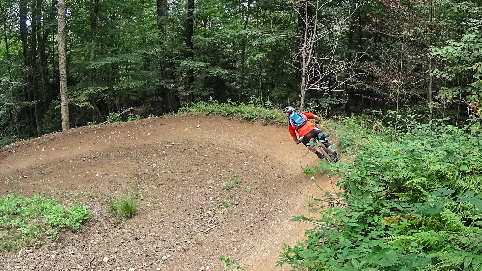 A mountain biker in an orange shirt navigates a dirt trail curving through a lush green forest. The scene captures the rider leaning into the turn, surrounded by foliage and trees. Snowshoe Bike Park mountain bike trail.