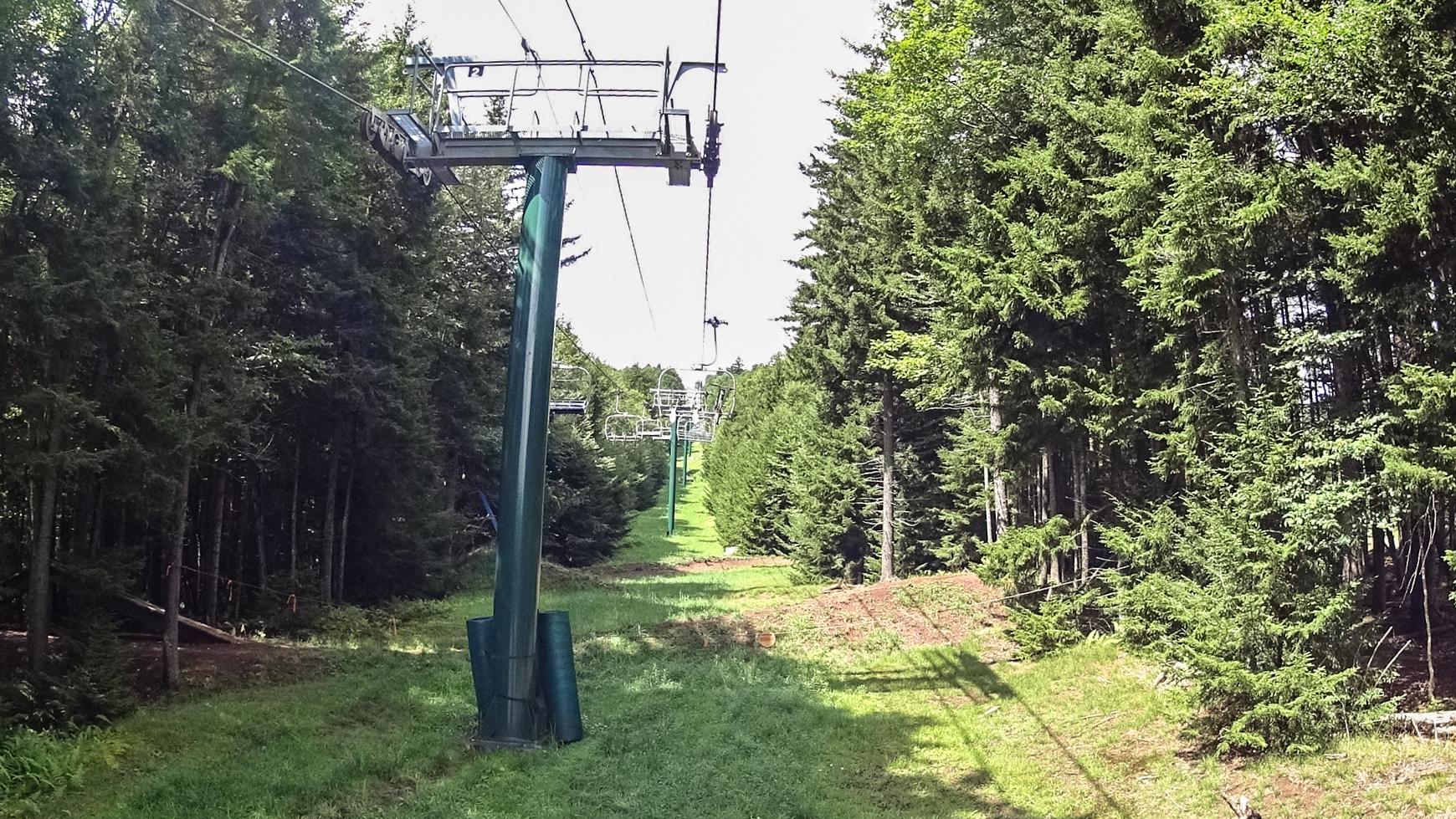 A scenic view of a ski lift running through lush green trees, with a clear path visible below and a bright sky above. The ski lift chairs are suspended above the ground, surrounded by dense forest on both sides. Snowshoe Bike Park mountain bike trail.