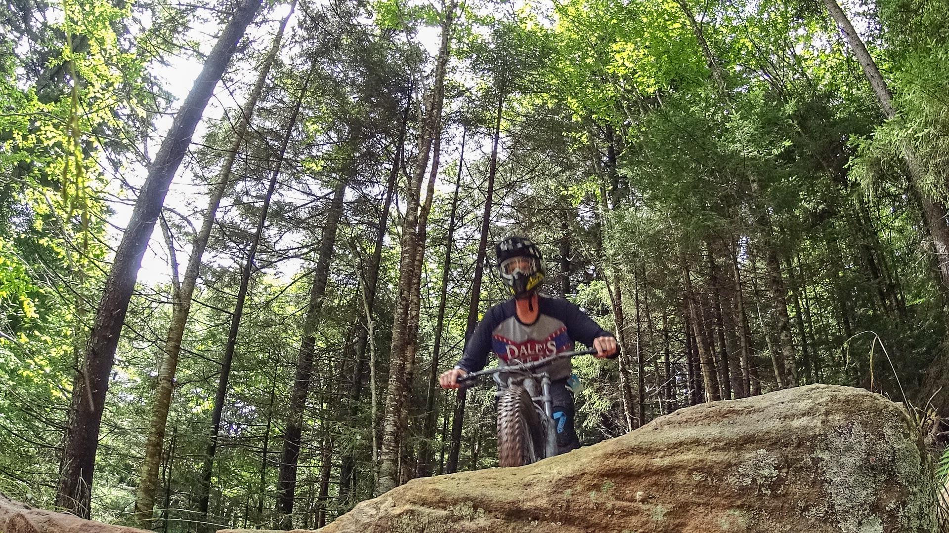 A mountain biker navigating over a large rock on a forest trail, surrounded by tall trees and greenery. The biker is wearing a helmet and protective gear, showcasing an adventurous moment in nature. Snowshoe Bike Park mountain bike trail.