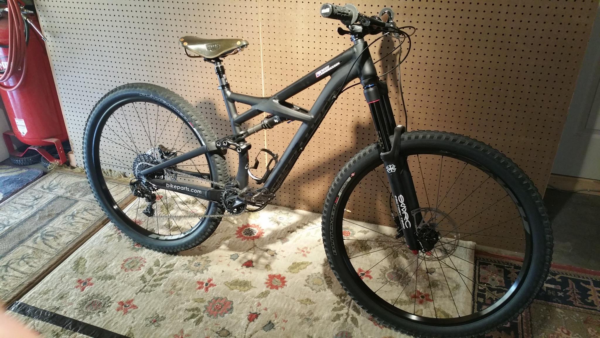 Specialized Enduro Comp 29: A black mountain bike leaning against a wooden pegboard, featuring a sleek frame, wide tires with tread, and front suspension. The bike is positioned on a patterned rug, with a glimpse of a red object visible in the background.