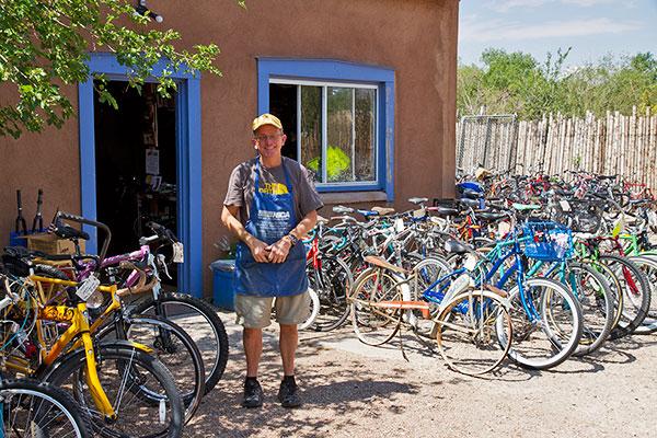 A smiling man in an apron stands in front of a bicycle shop, surrounded by a variety of bicycles parked outside. The shop has a warm, earthy exterior with a blue-framed window. Lush greenery is visible in the background, and the scene is bright and sunny.