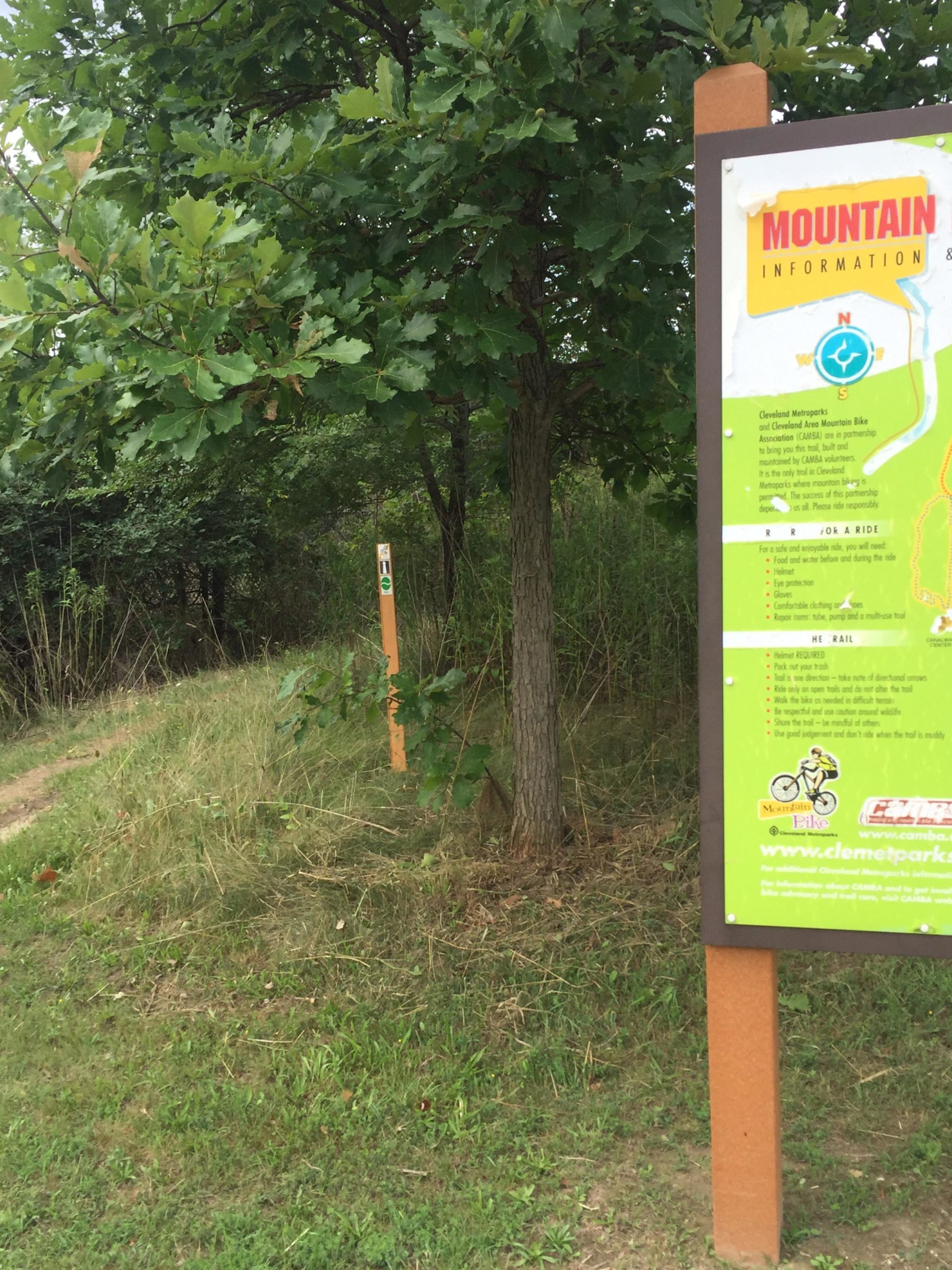A signpost for mountain biking information stands alongside a gravel path in a green, wooded area. The sign provides details for cyclists, including safety tips and guidelines, and features a map section. A tree with lush green leaves partially shades the area, and another marker is visible further down the path, indicating a designated trail. Ohio And Erie Canal mountain bike trail.