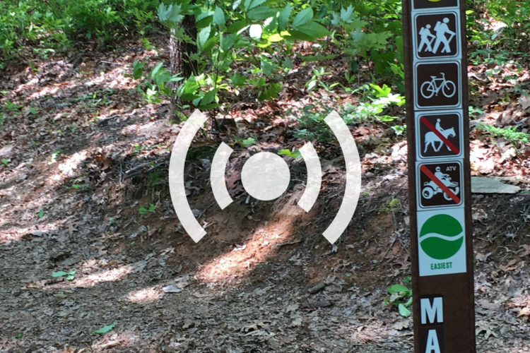 A trail sign in a wooded area, featuring symbols indicating allowed activities such as hiking and biking, and prohibiting horses and ATVs. The sign also includes information on the trail's difficulty level marked as "easiest." The background shows a natural landscape with leaf litter and greenery.