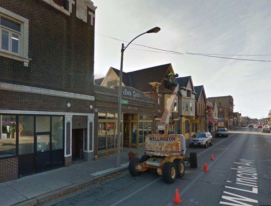 A street scene featuring a lift in front of a storefront called "Bent Lytle." A worker is seen on the lift, performing maintenance or construction at the top of the building. Several vehicles are parked along the street, with traffic cones placed nearby to mark the work area. The buildings feature a mix of architectural styles typical of a commercial district.