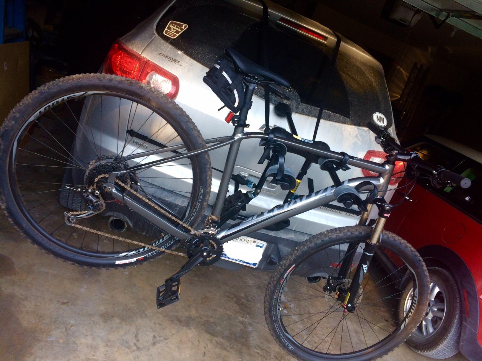 Specialized Rockhopper Comp 29: A mountain bike mounted on a car bike rack inside a garage. The bike has a gray frame and is equipped with thick tires, showing signs of dirt from outdoor use. In the background, there is a silver car with a dirty rear window and a red car partially visible. The garage floor is concrete and cluttered with various items.