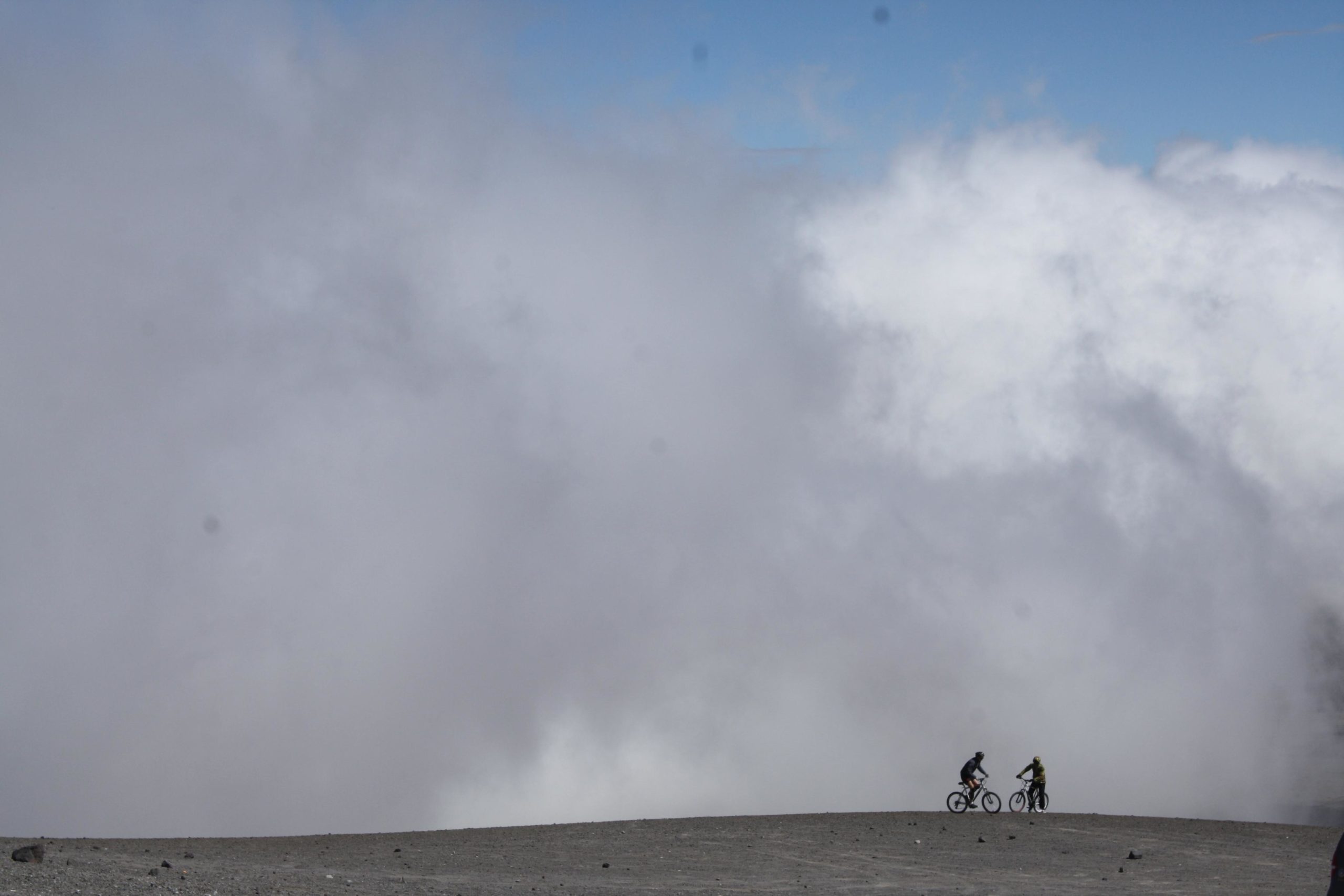 Two mountain bikers are silhouetted against a backdrop of gray clouds and a blue sky, riding on a vast, barren landscape with rocky terrain. Cotopaxi Volcano National Park mountain bike trail.
