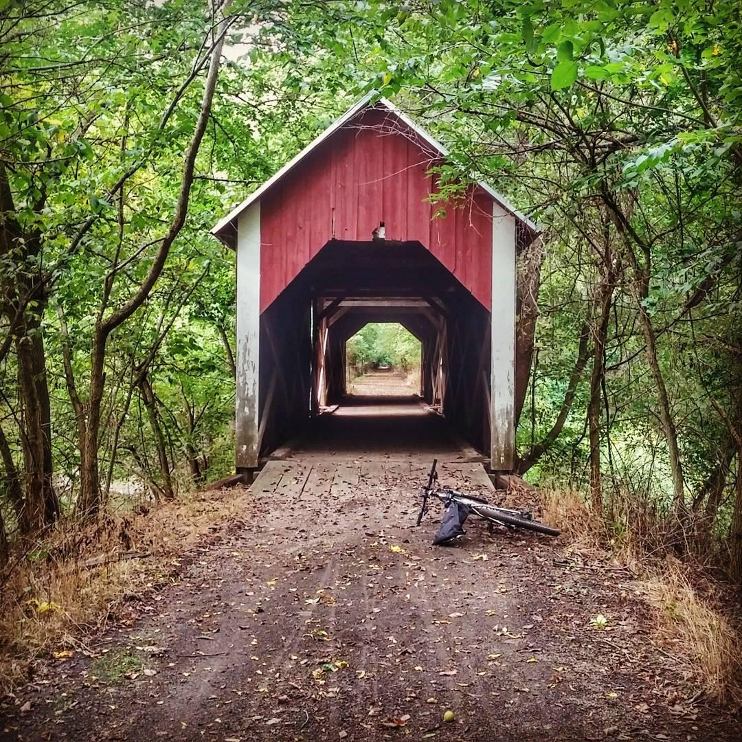 A rustic covered bridge surrounded by lush greenery, with a dirt path leading through it. On one side of the path, there is a bicycle resting against a wooden support, and fallen leaves cover the ground. The bridge features a red exterior and white accents, creating a charming atmosphere. Cinder Path mountain bike trail.