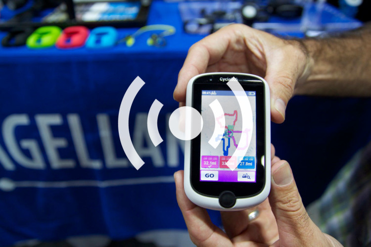 A person holding a cycling device with a colorful map display, featuring GPS navigation and various cycling metrics. In the background, multiple cycling accessories are partially visible on a blue table.