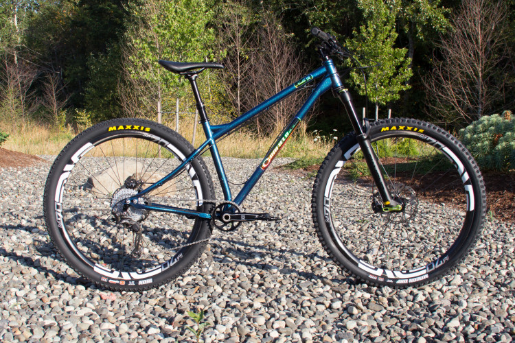 A blue mountain bike with a sleek design, featuring thick Maxxis tires, positioned on a gravel surface surrounded by greenery. The bike has a sturdy frame and front suspension, ready for adventure.