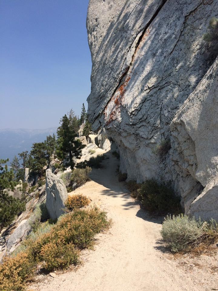 A winding dirt path leading through rocky terrain, flanked by large boulders and patches of greenery, set against a clear blue sky. Flume Trail mountain bike trail.