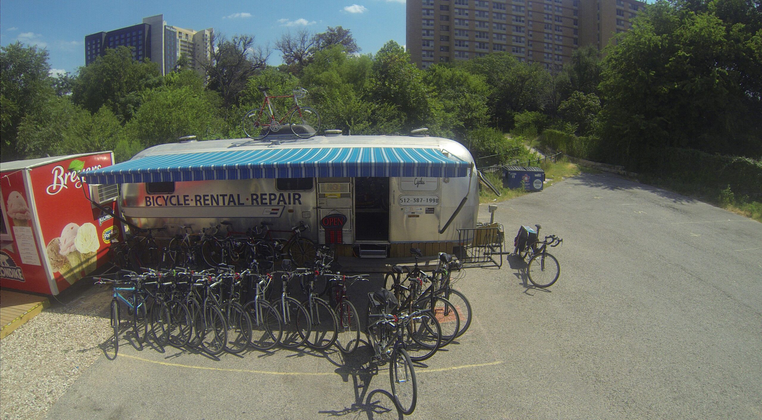 A retro-style Airstream trailer with a blue and white striped awning, serving as a bicycle rental and repair shop, surrounded by several parked bicycles. In the background, a green landscape and residential buildings are visible under a clear blue sky. An ice cream truck is nearby, adding to the vibrant outdoor scene.
