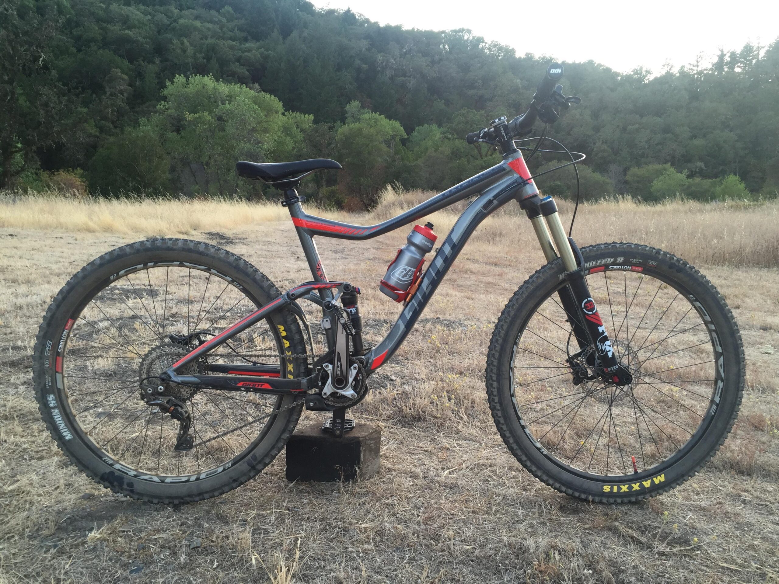 Giant Giant Trance 27.5 2: A mountain bike with a grey and red frame is positioned on a dry, grassy terrain, surrounded by trees in the background. The bike features thick tires, a water bottle attached to the frame, and is standing on a small block. The setting suggests an outdoor, adventure-oriented environment.