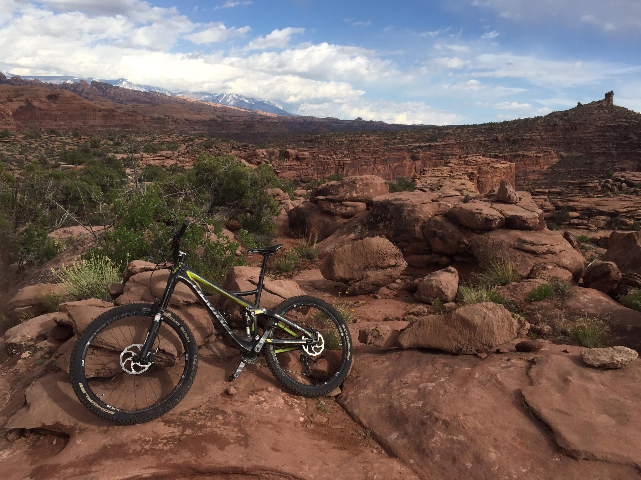 Devinci Troy Carbon RR: A mountain bike leaning against a rock formation in a rugged desert landscape, with distant mountains partially covered in snow under a cloudy sky.