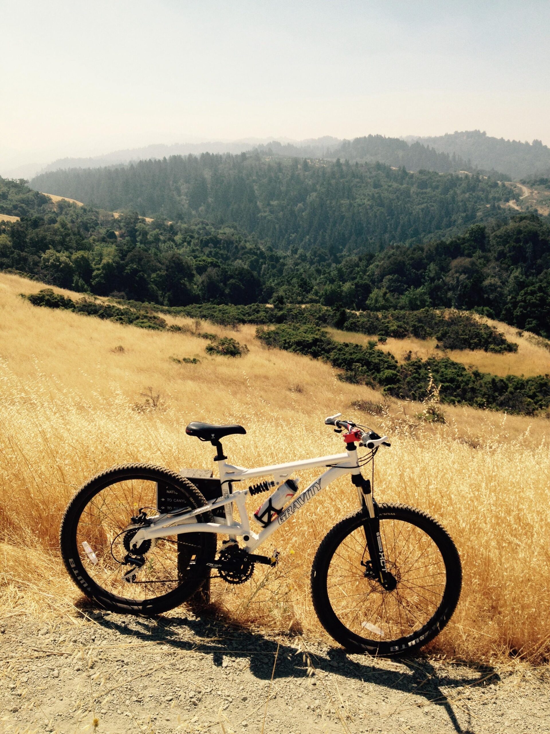 A mountain bike parked on a dirt path overlooking a scenic mountain landscape with rolling hills and trees. The foreground features dry grass, while the background shows a hazy view of distant mountains. Monte Bello / Rancho San Antonio mountain bike trail.