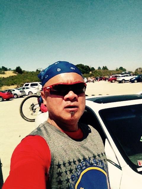 A person wearing sunglasses and a blue bandana stands next to a white car in a parking area. In the background, there are several parked cars and green hills under a clear blue sky. The individual is dressed in a gray tank top and red long-sleeve shirt, appearing to be outdoors, possibly preparing for a biking activity. Monte Bello / Rancho San Antonio mountain bike trail.