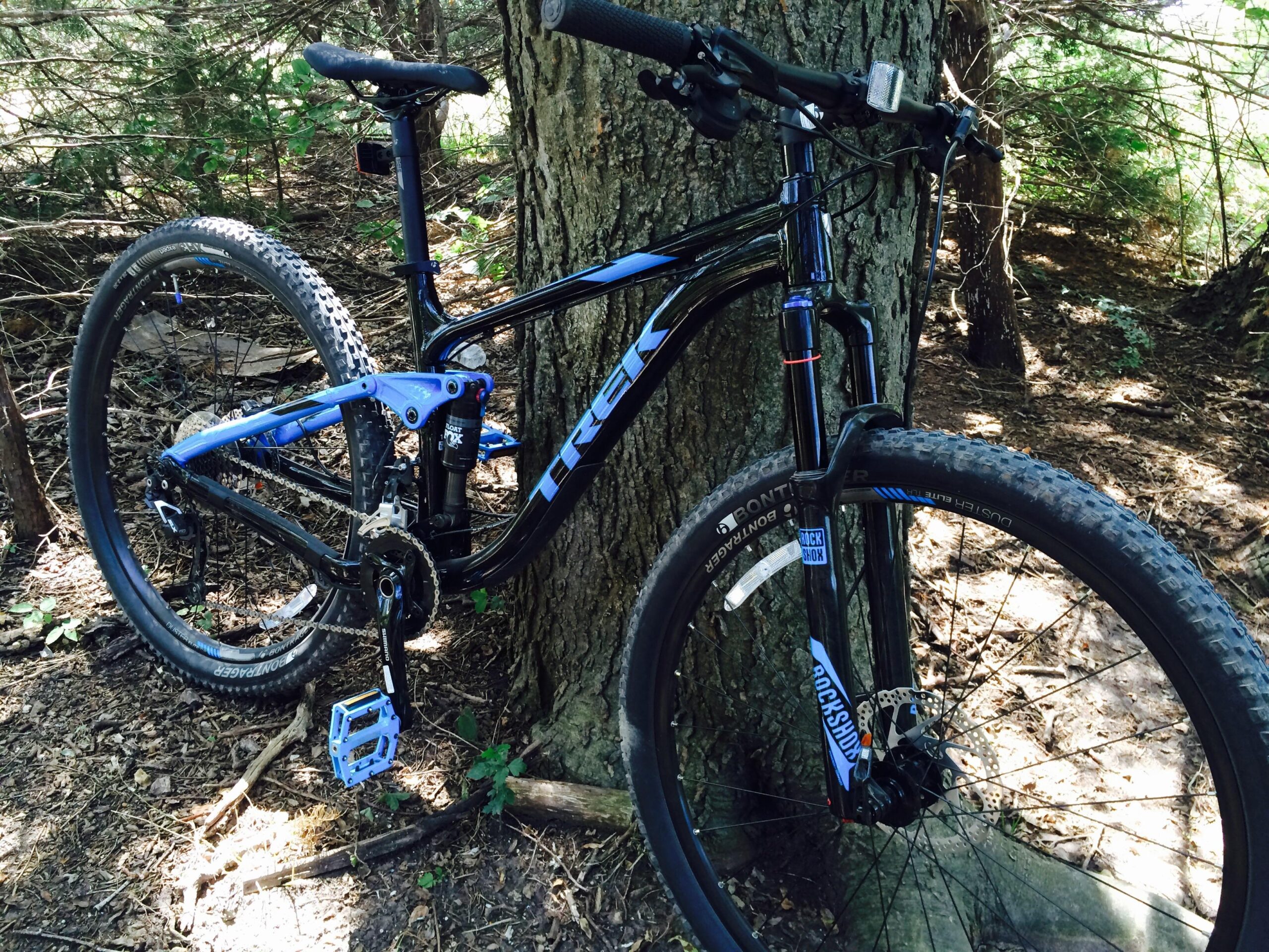 Trek Fuel EX 7 29: A black and blue mountain bike resting against a tree in a wooded area, featuring knobby tires and a suspension system.