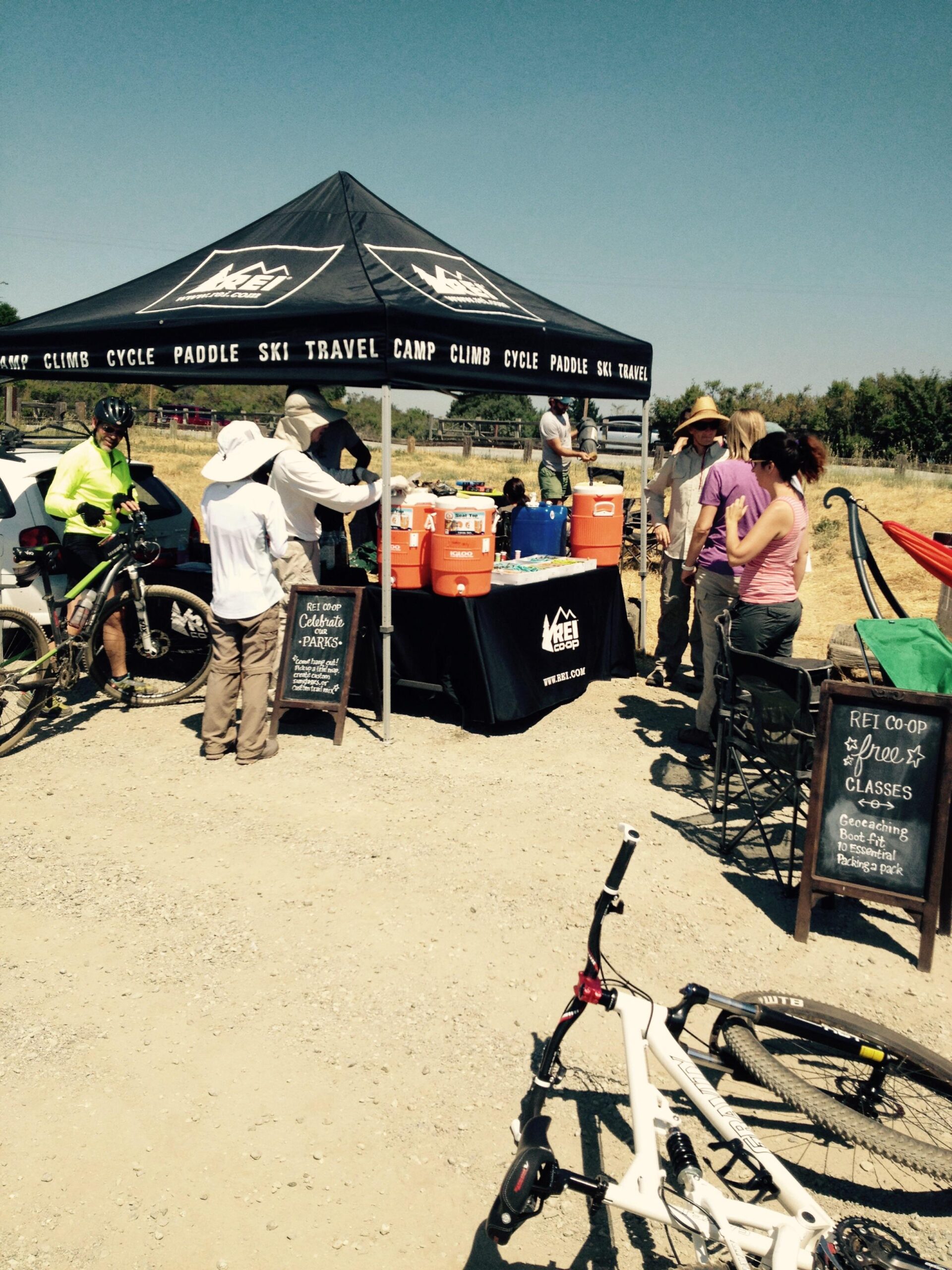 A sunny outdoor scene featuring a REI Co-op tent with a banner displaying outdoor activities like camping, climbing, and cycling. Several people are gathered near the tent, engaging with the staff and enjoying refreshments from large coolers. Bicycles are leaning nearby, and a chalkboard sign advertises free classes. The background shows a clear blue sky and a grassy area. Monte Bello / Rancho San Antonio mountain bike trail.