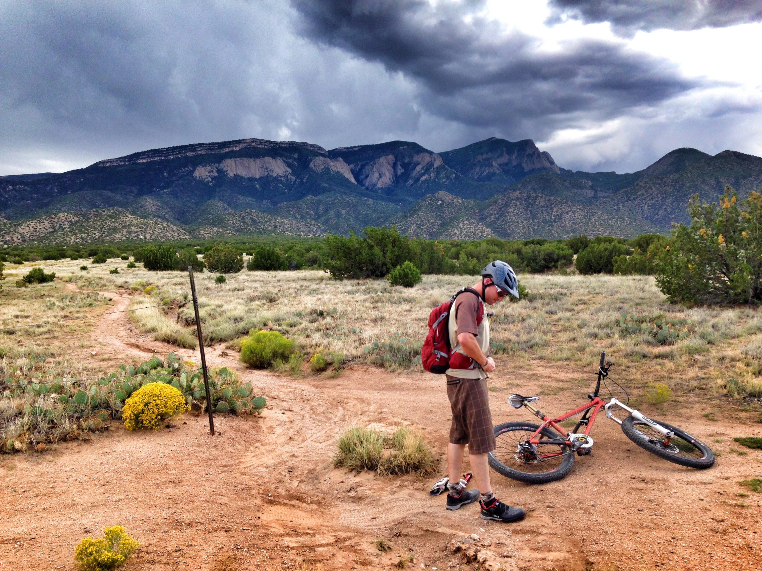 A mountain biker inspects their bike in a grassy area with a dirt trail, set against a backdrop of dark, cloudy skies and distant mountains. The biker is wearing a helmet and a backpack, standing beside an orange mountain bike that is laid on its side. Cacti and shrubs are scattered around, creating a natural landscape. Up & Up mountain bike trail.