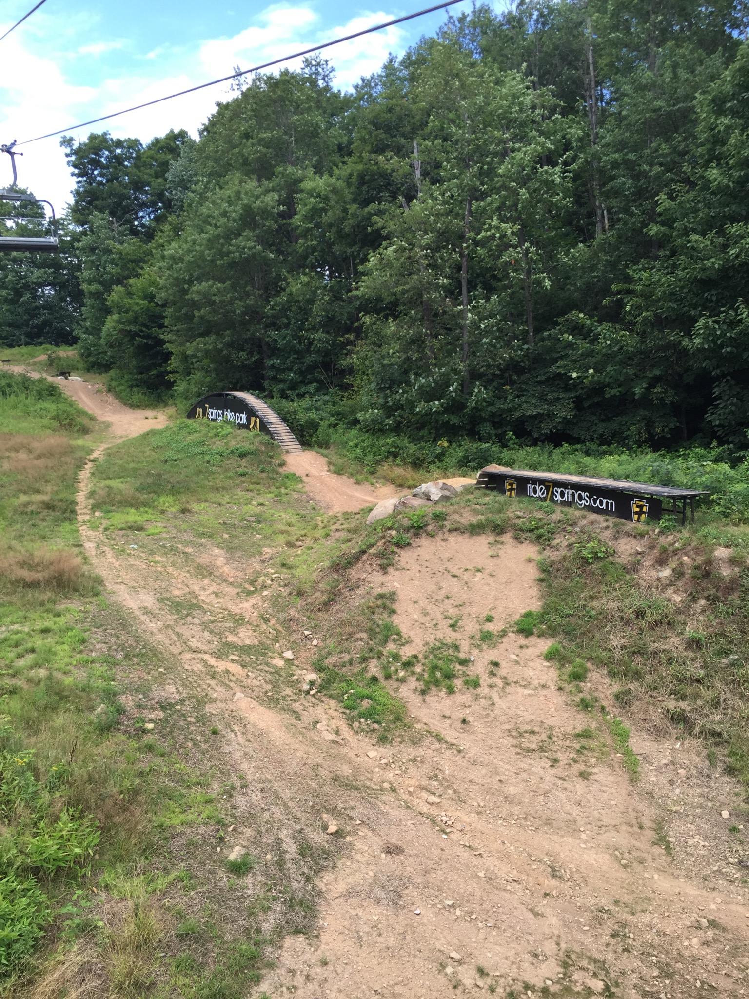 Alt text: A scenic view of a winding dirt biking trail surrounded by green trees, featuring a wooden ramp labeled with the website "ride2sprints.com." A ski lift can be seen in the upper left corner against a blue sky. Seven Springs mountain bike trail.