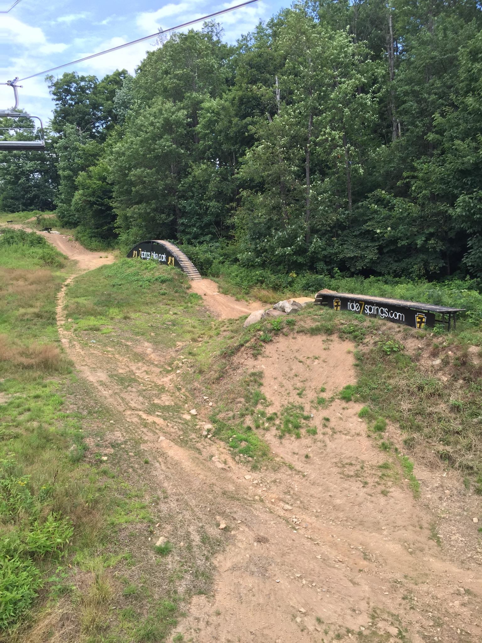 A scenic view of a mountain bike trail featuring a wooden ramp in the foreground. Lush green trees line the background, and the path is marked with signage promoting a bike park. The sky is partly cloudy, hinting at a pleasant day for outdoor activities. Seven Springs mountain bike trail.