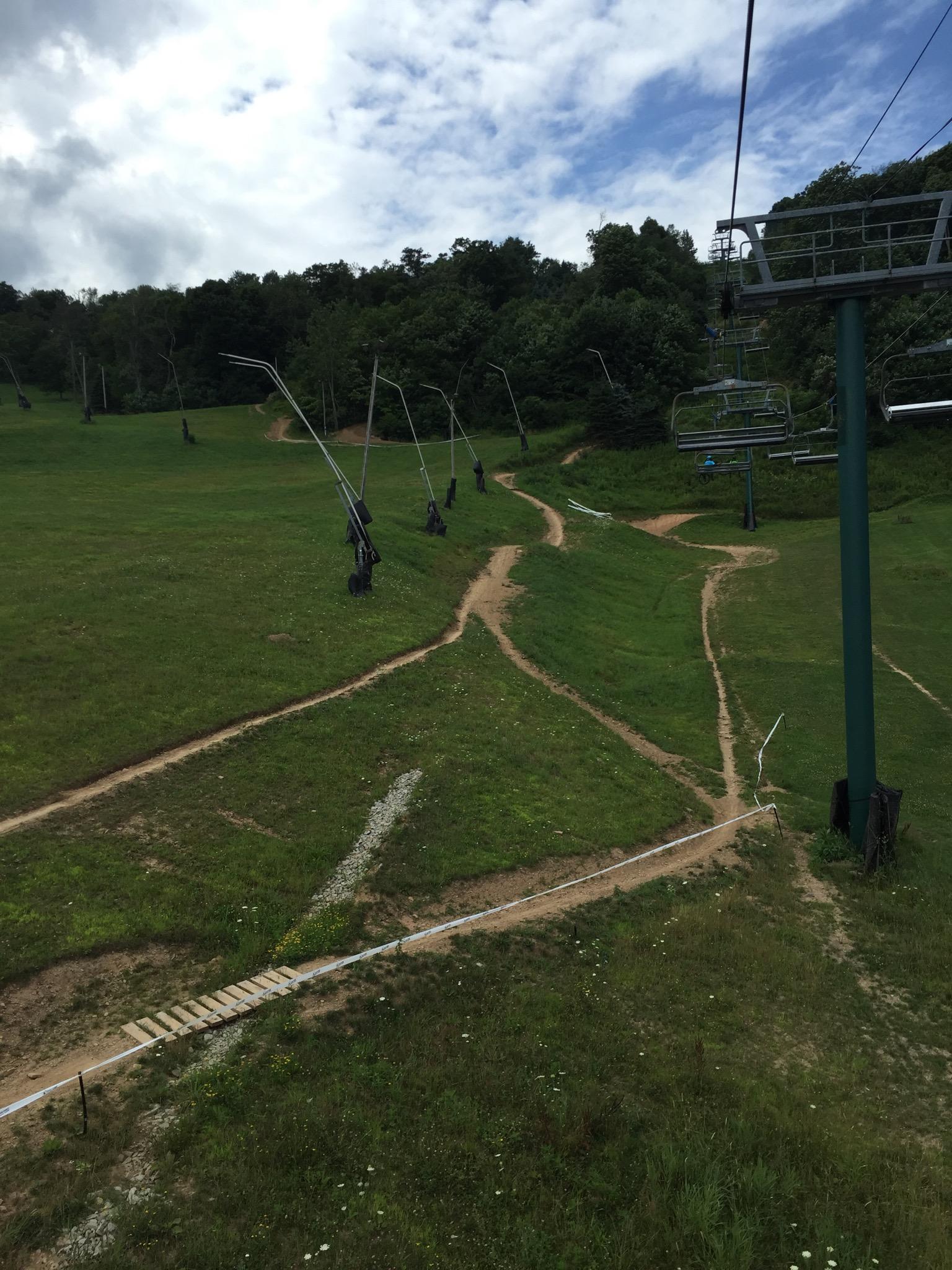 A scenic view from a ski lift overlooking a lush green hillside, with a few dirt paths winding through the grass. In the foreground, part of the ski lift structure is visible, while the background features trees and a cloudy sky. Seven Springs mountain bike trail.