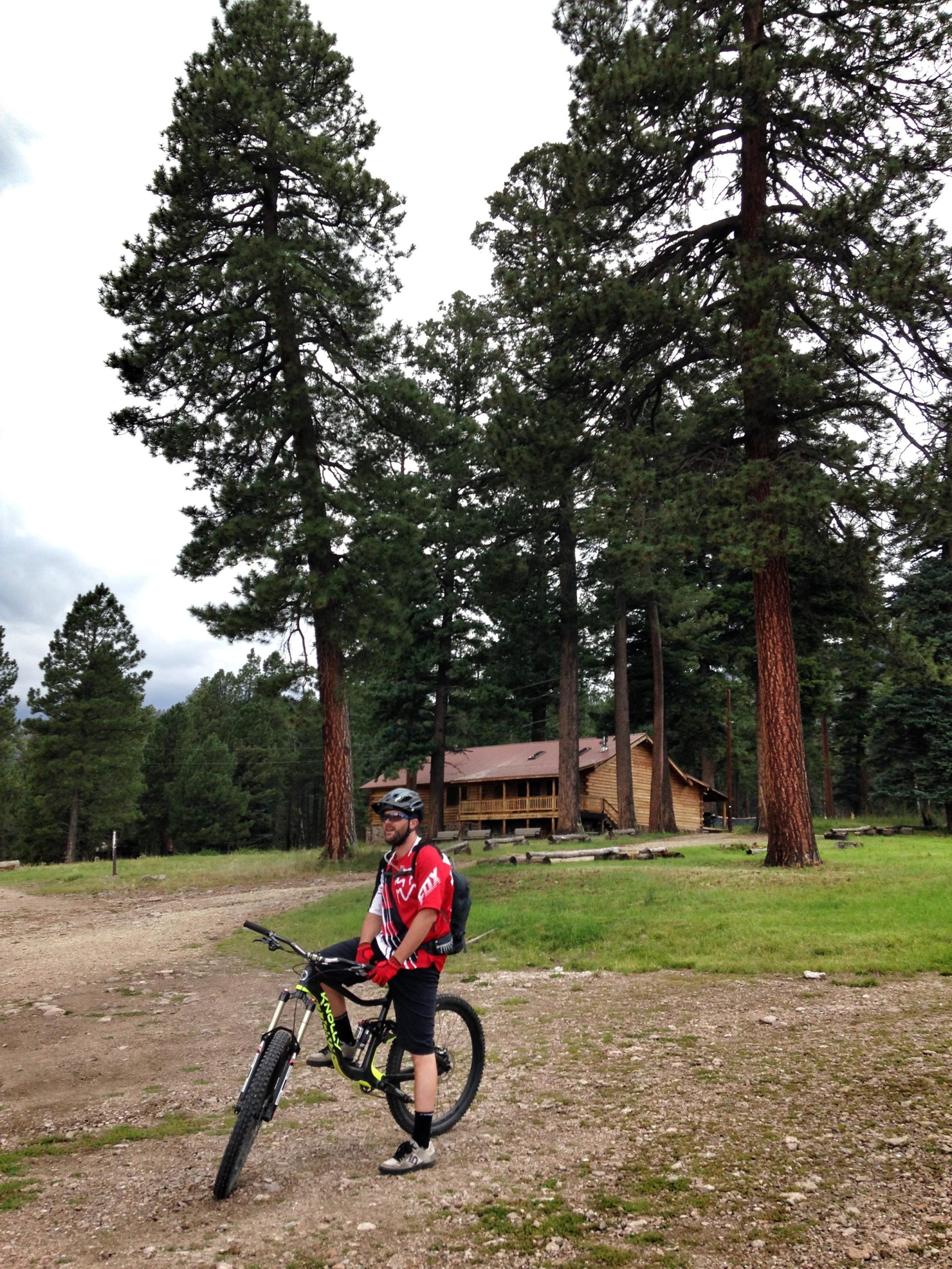 A mountain biker wearing a red jersey and black shorts stands next to a bicycle on a gravel path. Tall green pine trees surround the area, and a wooden cabin is visible in the background, partially hidden by the trees. The sky is overcast, indicating a cool, cloudy day. Valles Caldera Trails mountain bike trail.