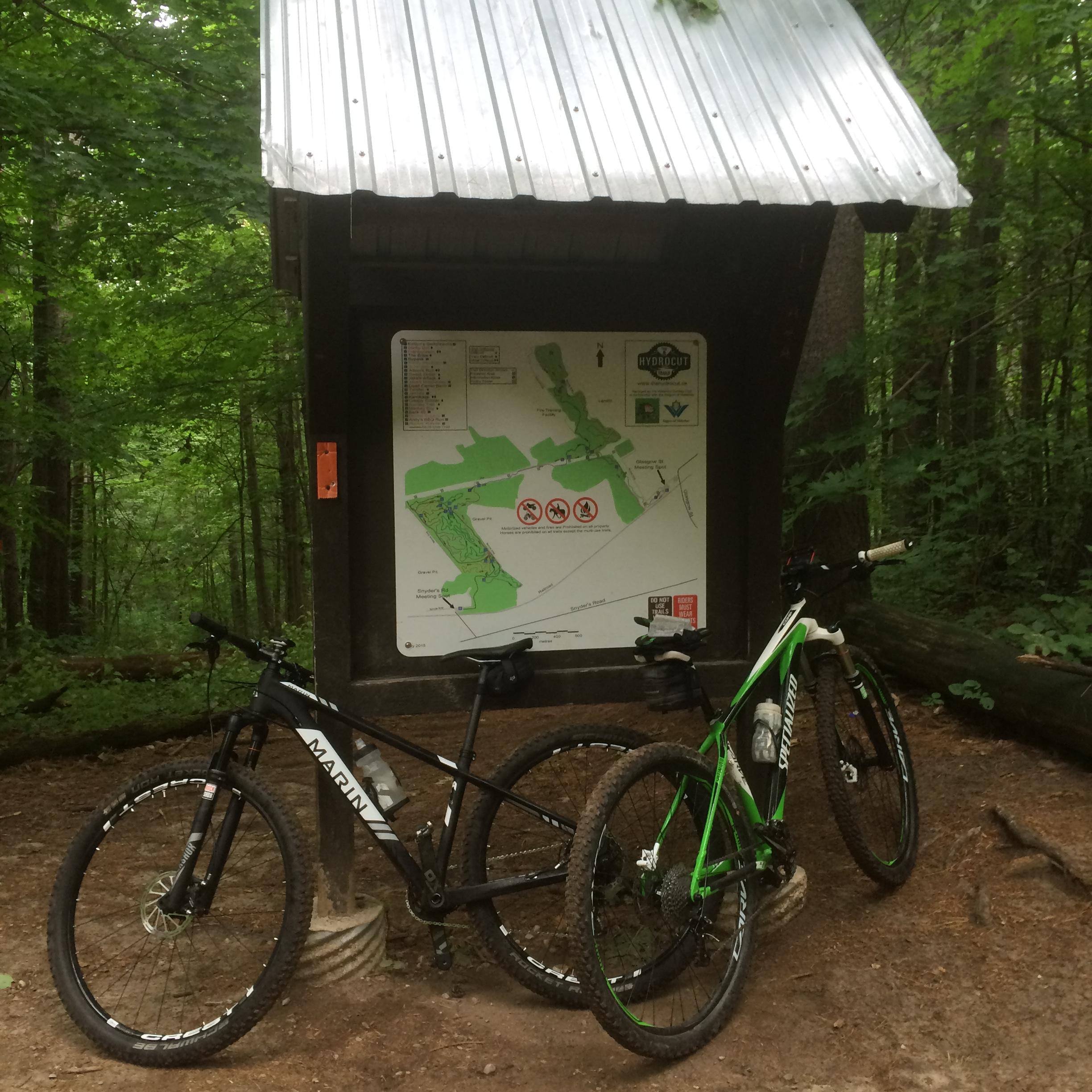 A pair of mountain bikes, one black and one green, leaning against a wooden structure with a metal roof. The structure features a map highlighting a trail area surrounded by lush green trees in a forested setting. Hydrocut mountain bike trail.