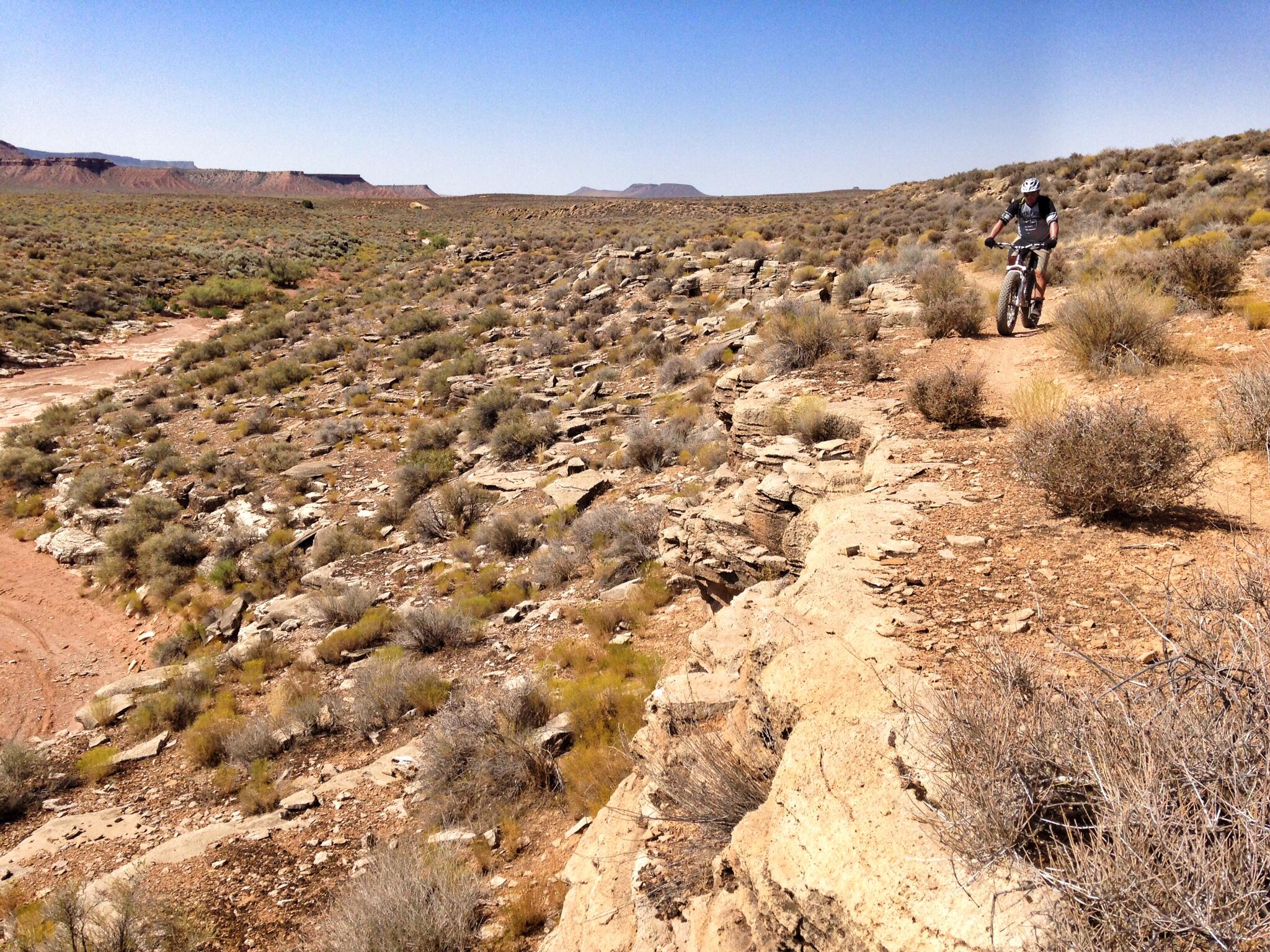 A person riding a mountain bike along a rocky trail in a desert landscape, with sparse vegetation and distant plateaus under a clear blue sky. J.E.M. Trail mountain bike trail.