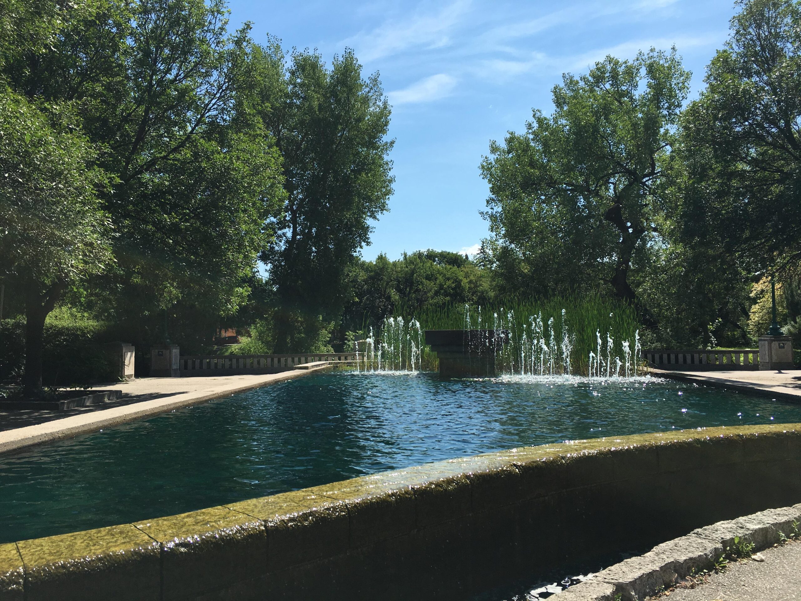A sunlit view of a tranquil park featuring a clear blue pond with decorative fountains. Surrounding the pond are lush green trees and well-maintained pathways, creating a serene and inviting atmosphere. Whittier Park & Parker Lands & Churchill Dr mountain bike trail.